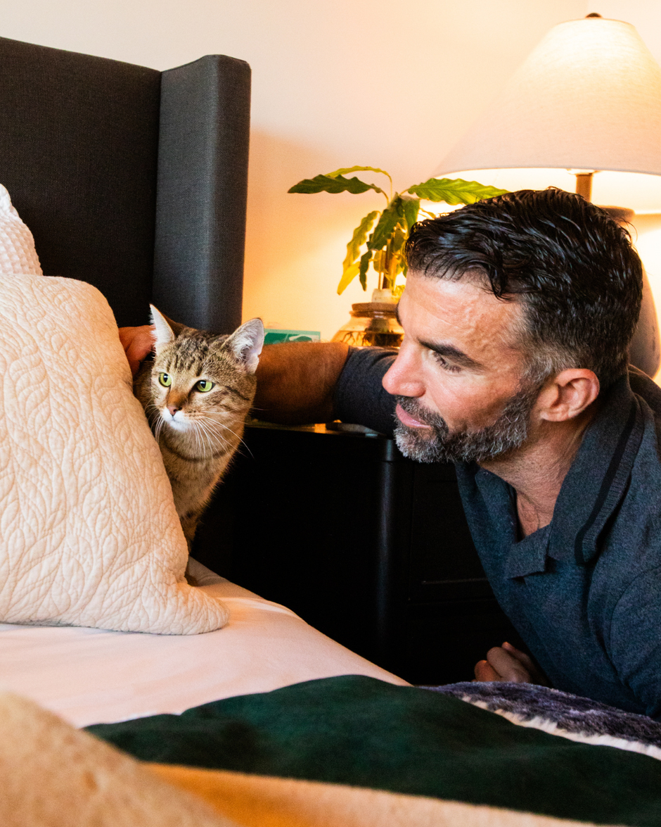 Man sitting on a bed with a cat looking at him, in a cozy bedroom setting.
